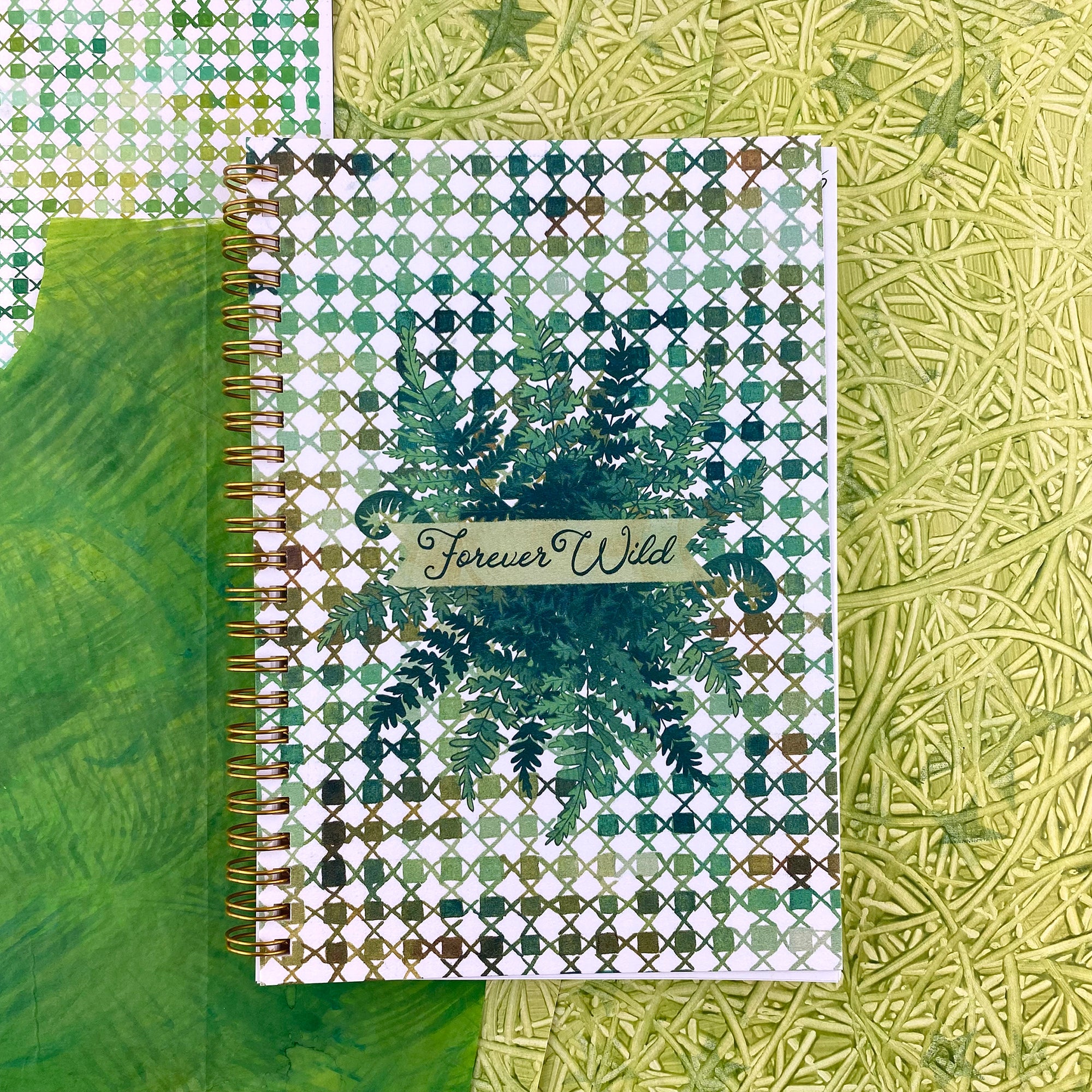 Spiral Bound notebook with a handpainted checkered cover , a grouping of ferns in the center with a banner that says Forever Wild in a hand drawn script font. Very outdoorsy. Using all the green colors. 